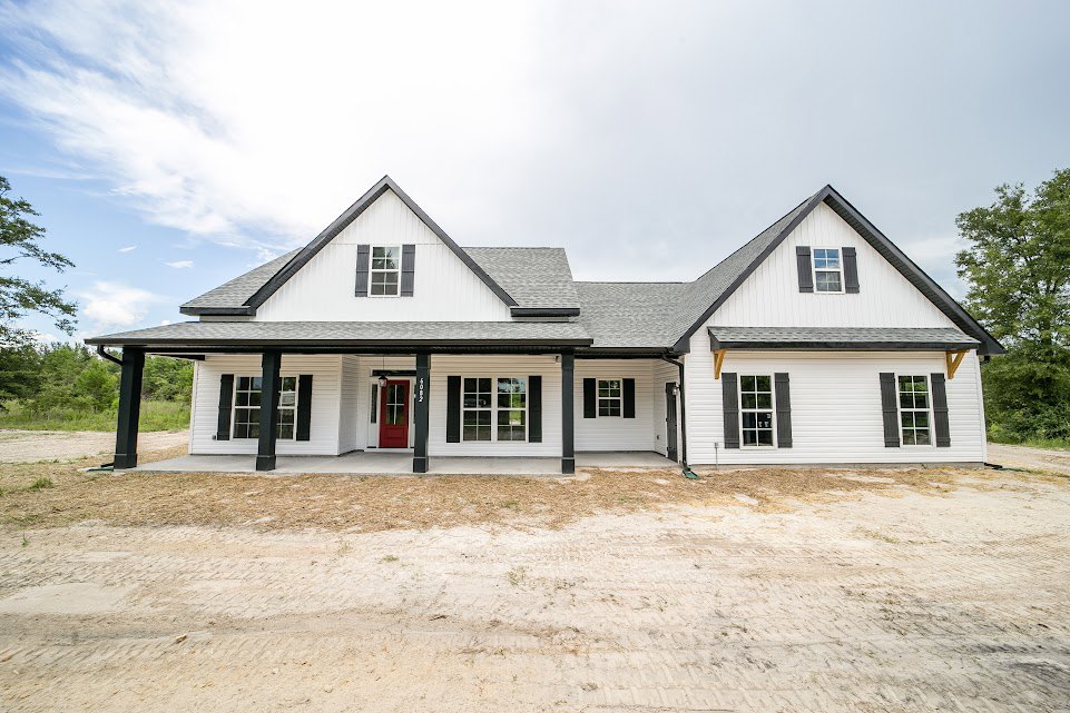 White siding house with black trim, red front door, white-framed windows, dirt yard, tree and cloudy sky in background