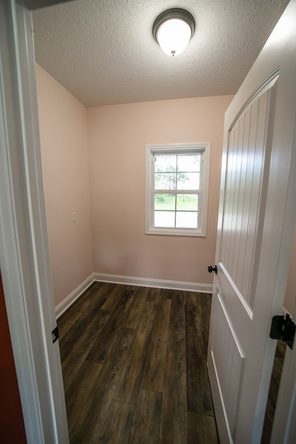 Wood floor with white trim, white paneled walls, single window overlooking a field of trees, white door, ceiling light fixture with exposed bulb