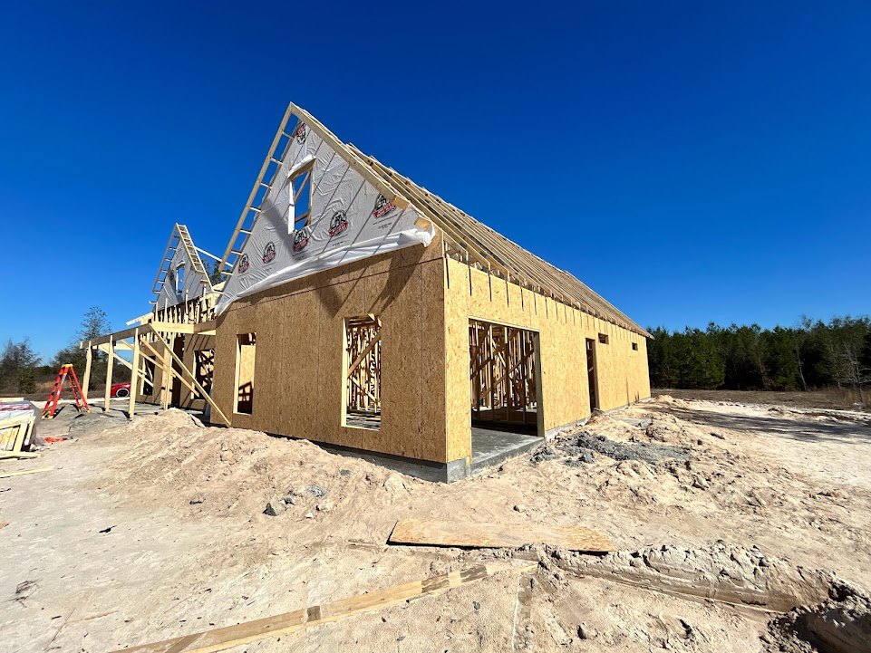 Wood-framed house under construction on sandy ground, partially completed roof, red ladder leaning against concrete foundation, surrounding trees in background