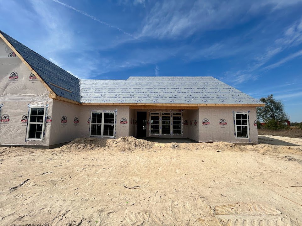 Two-story house under construction with white-framed multi-pane windows, sandy dirt pile in front, and clear blue sky overhead