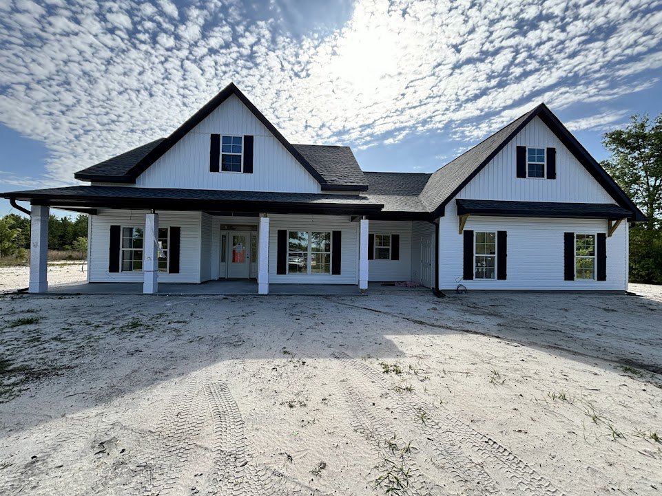 Two-story house with black roof, large sandy driveway showing tire tracks, white front door with sign, window reflecting parked car, cloudy sky overhead, trees at property edge