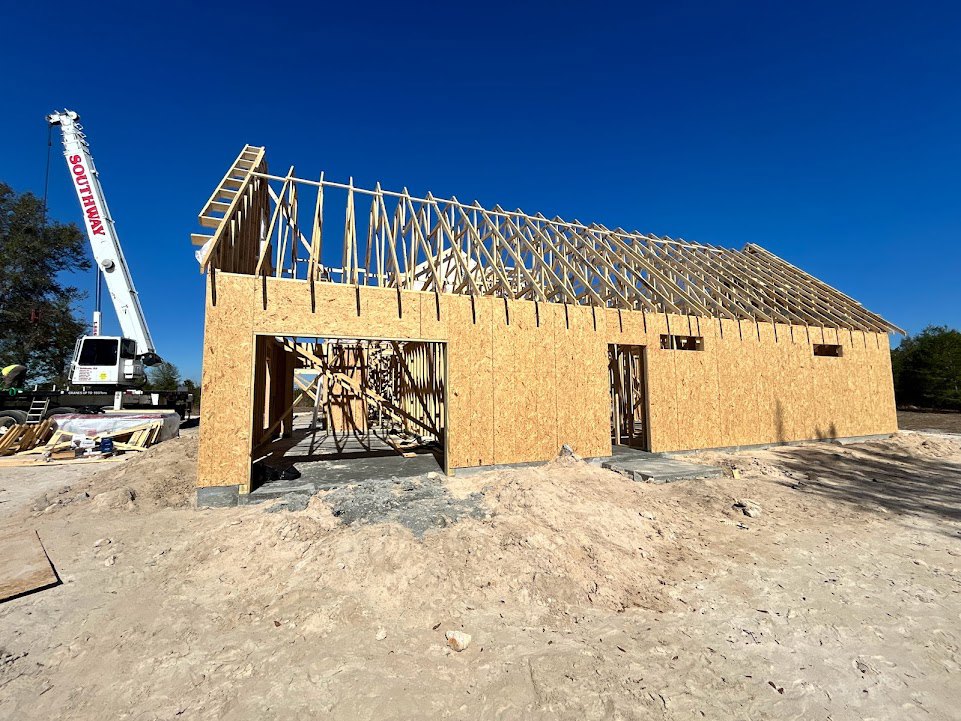 Wood-framed roof under construction, white crane with red lettering beside unfinished exterior walls, pile of dirt and white truck in foreground, clear blue sky overhead