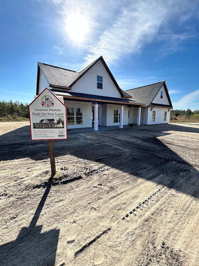 White two-story house with columned porch, red sign on post in front yard, large windows, cloudy sky overhead