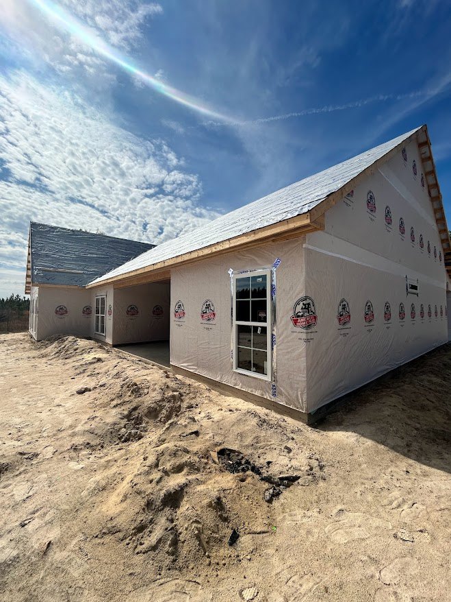Framed house under construction with exposed plywood walls, glass window installed, pile of dirt in foreground, white building nearby, roof partially covered with snow, blue sky