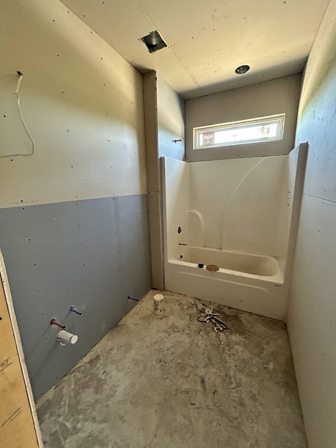 Modern bathroom with white bathtub, glass shower enclosure, large window, plaster walls, concrete floor, and metal wall-mounted light fixture