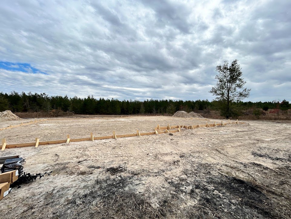 Dirt field bordered by a wooden fence, leafy tree in foreground, cloudy sky overhead, scattered trees in background