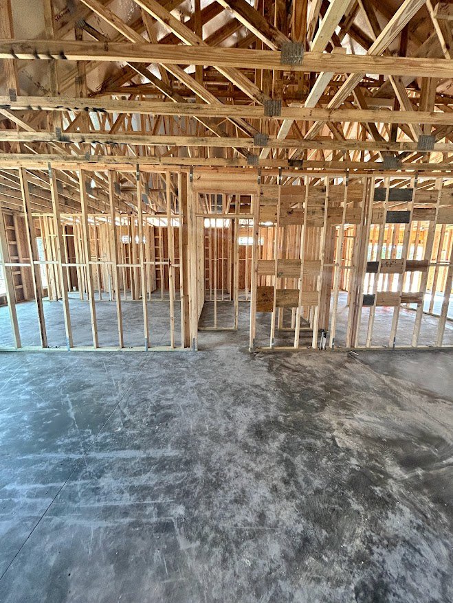 Wood-framed house under construction with exposed beams, concrete floor, wooden roof structure, and window in unfinished room