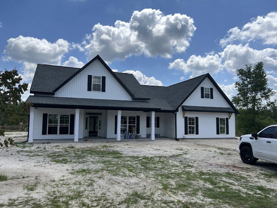 White house with black roof, white truck parked on dirt road, yellow rope on bare ground, green-leaved tree, blue sky with clouds