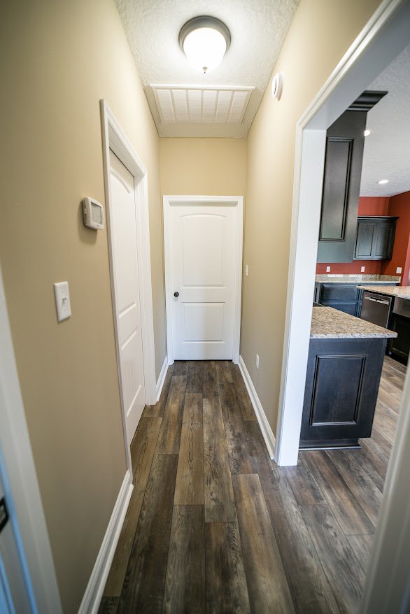 Hallway with light wood flooring, white door with black knob, black cabinet topped with marble, red accent wall, white ceiling with exposed beams