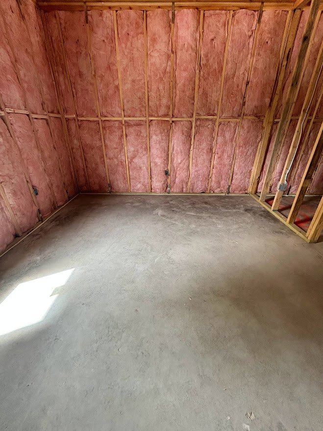 Concrete floor and white wall in a room with exposed wooden ceiling beams, sunlight illuminating a section of the wall, curtain partially visible.