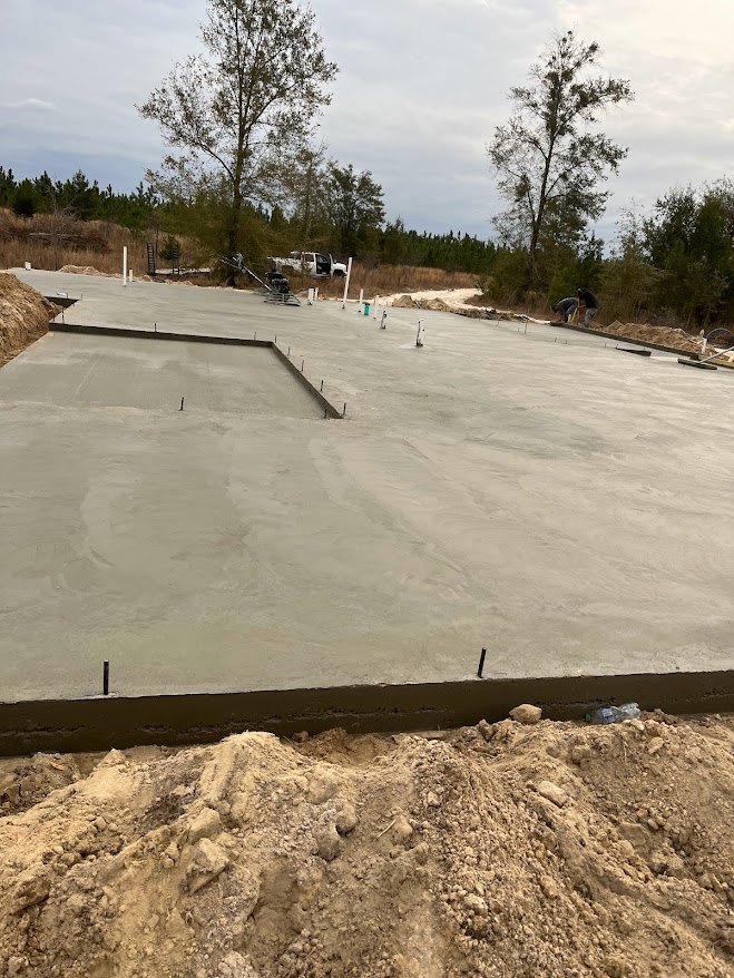 Freshly poured concrete slab with a square opening in the center, surrounded by piles of dirt and bordered by trees under a cloudy sky