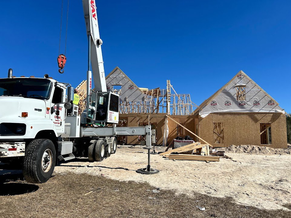 Crane truck parked beside a partially built house with exposed framing, triangular roof, and ladder; construction materials and equipment visible on site.