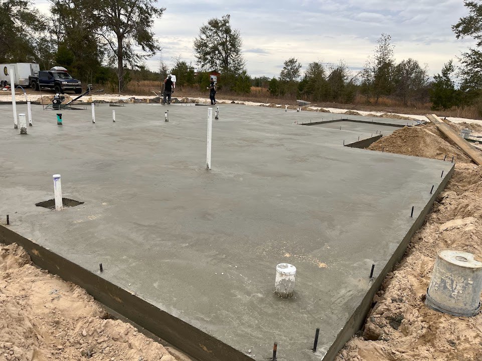 Concrete foundation slab with multiple metal poles protruding, surrounded by dirt and construction materials; blue and white trucks parked nearby under a partly cloudy sky.