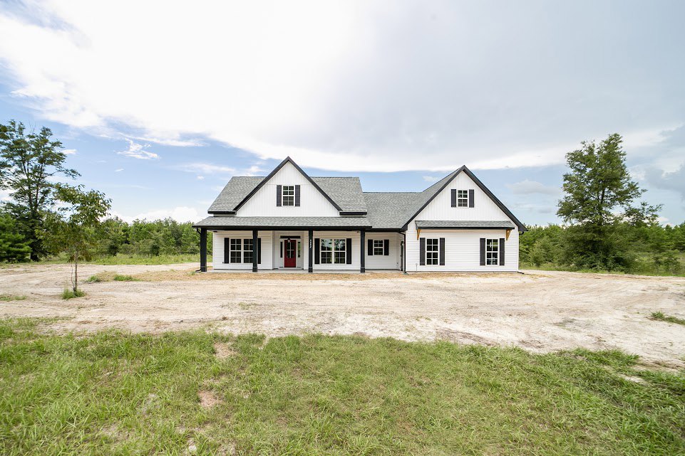 White house with red doors, gray roof, dirt road bordered by grass, mature tree with green leaves, blue sky and scattered clouds in background