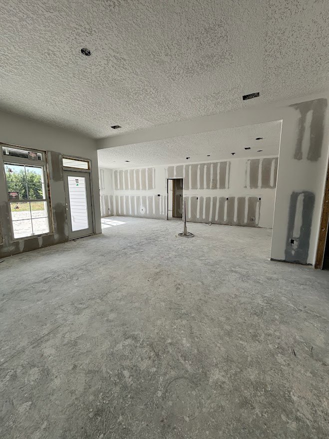 White-walled room with a door, large windows overlooking trees, dark-painted accent on one wall, central support pole, and a blurry shadow cast on the floor.