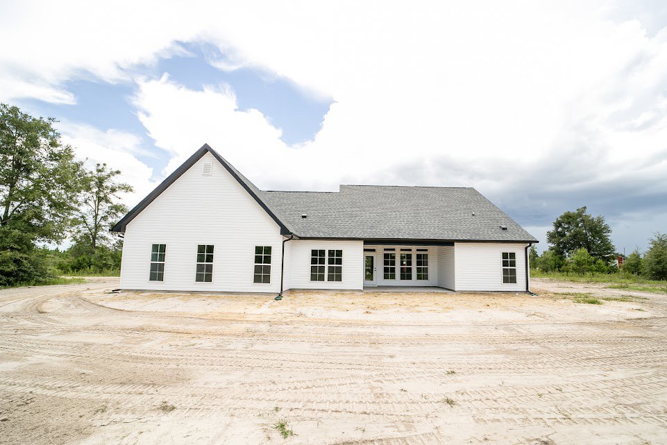 White house with black roof, tire tracks in sandy dirt yard, single tree nearby, blue sky with scattered clouds