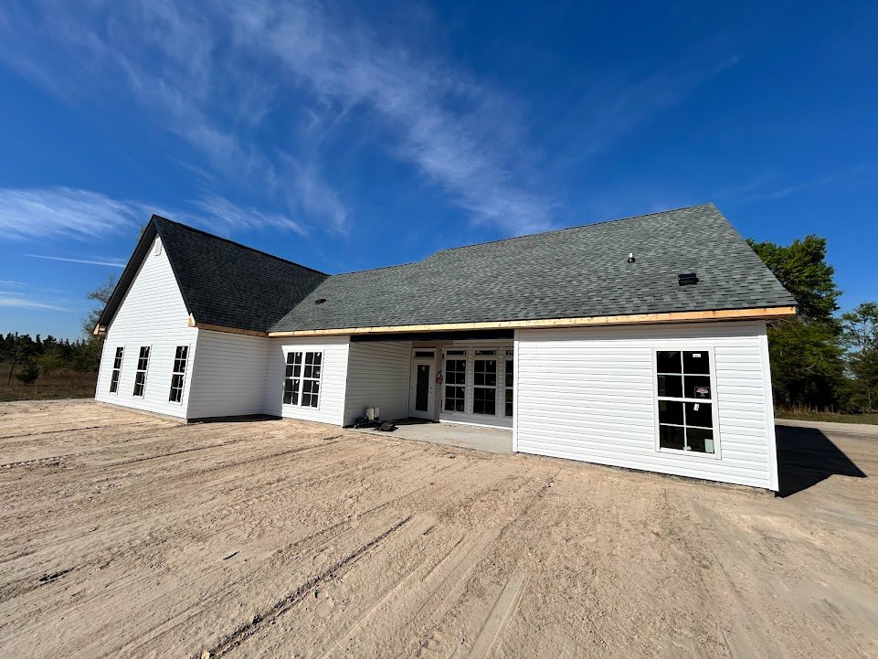 Partially built house with exposed framing, white garage door, stickered window, dirt yard with tire tracks, and clear blue sky overhead