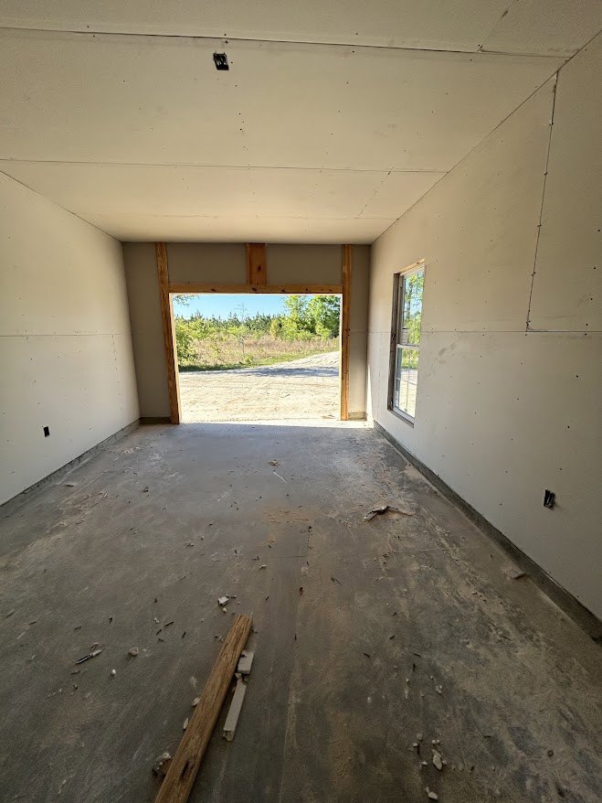 Concrete floor with exposed wood beam, white ceiling featuring a cutout, large window and doorway overlooking dirt road and trees, unfinished wall with wood and tile materials