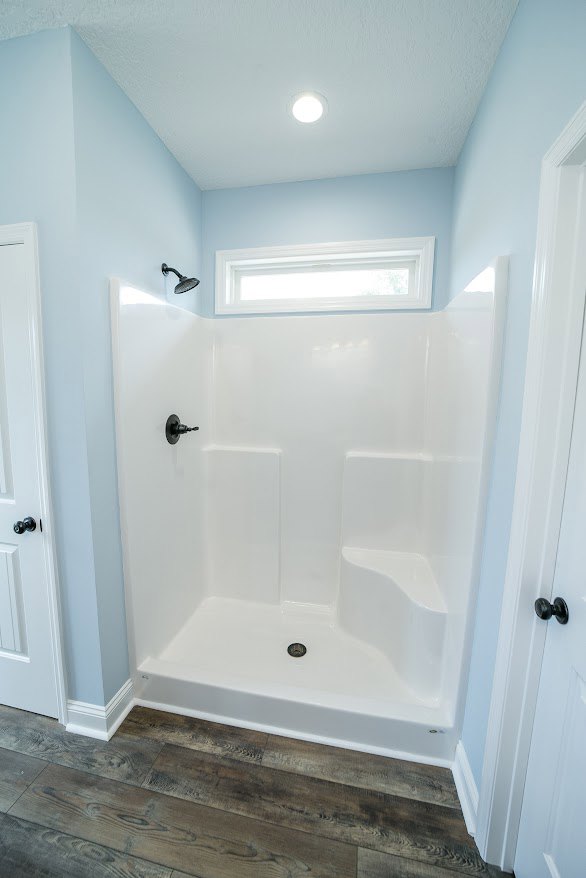 White framed shower with black handle, wood flooring with white baseboard, white door with square trim, plaster walls in modern bathroom.