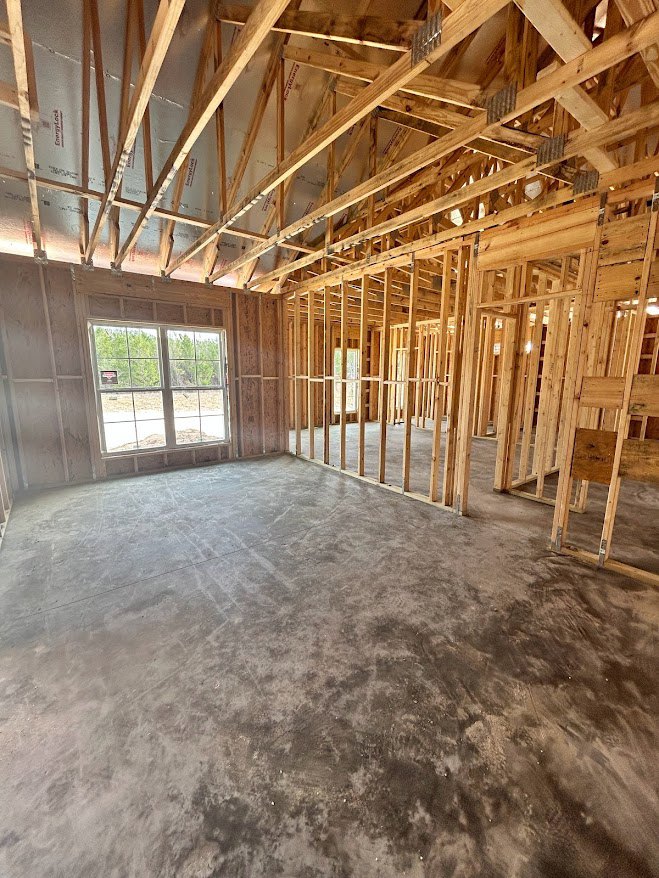 Wood-framed room under construction with exposed ceiling beams, multi-pane window, unfinished walls, and visible floor planks