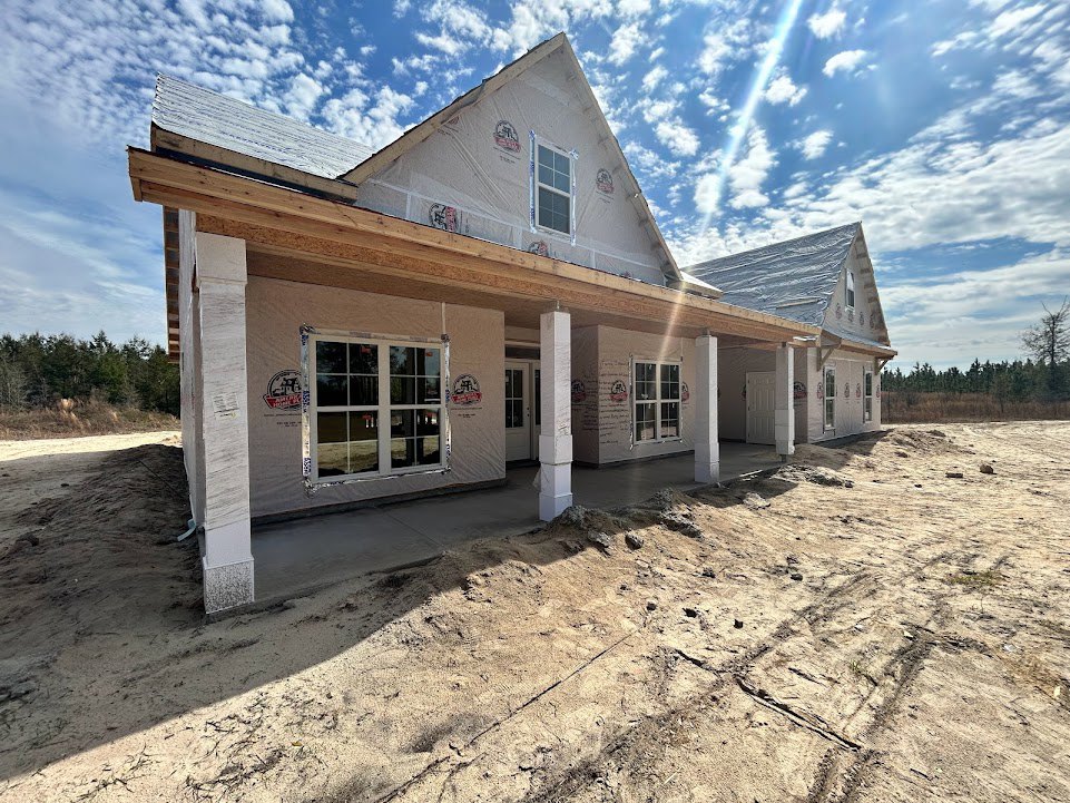 Framed house under construction with exposed wood, multi-pane window, dirt pile in foreground, and blue sky with scattered clouds