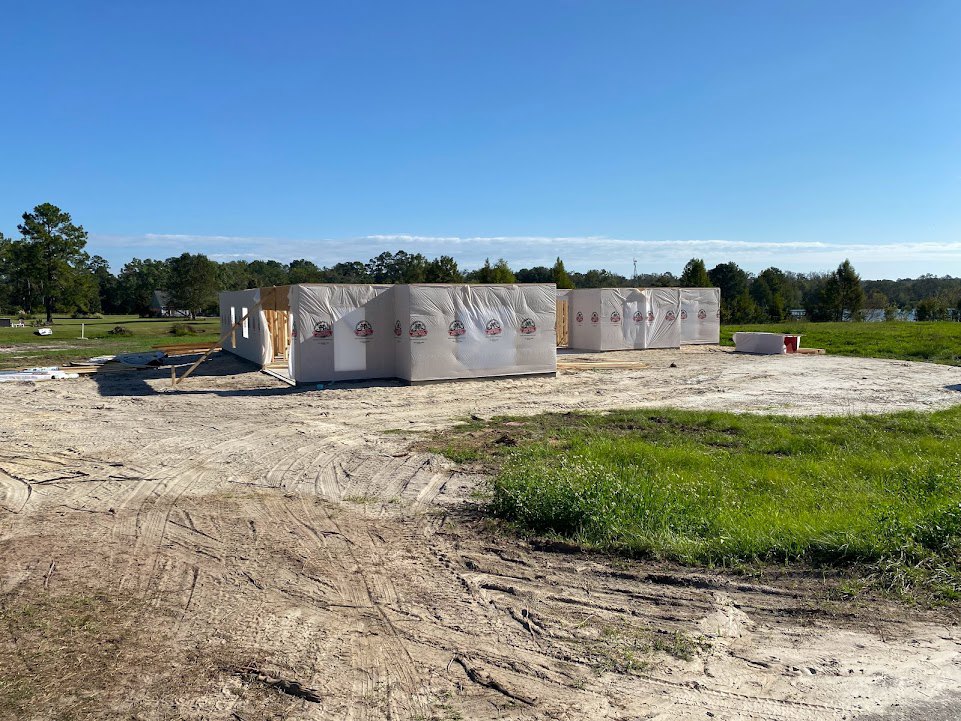 Wood-framed custom home under construction with exposed beams, surrounded by grassy lot, mature trees, and blue sky with scattered clouds
