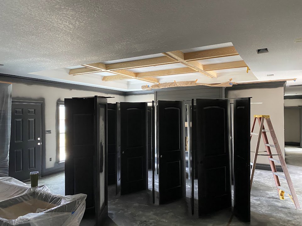 Room under construction with multiple black and white doors, silver handles, exposed beams, ladder on the floor, and plastic-wrapped sink.