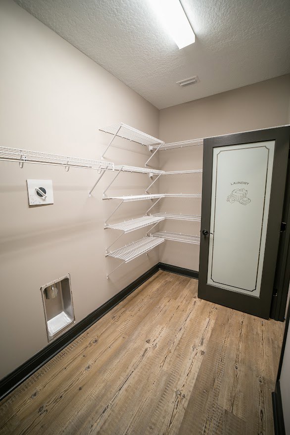 Bathroom with wood flooring, built-in white shelves, plaster walls, and a door featuring a framed picture; silver urinal with tissue paper visible.