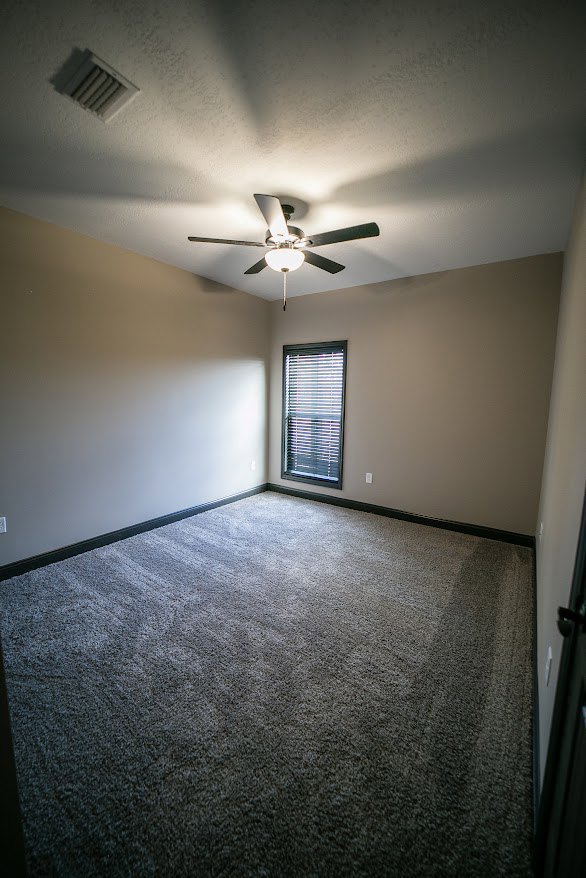 Carpeted room with white plaster walls, ceiling fan with light fixture, ceiling vent, and window fitted with horizontal blinds