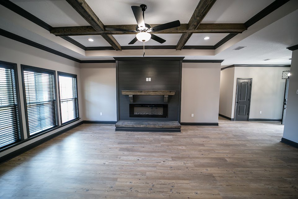 Living room with wood flooring, white plaster walls, central fireplace topped by a wood mantel, ceiling fan with light fixture, grey door with black handle, window fitted with