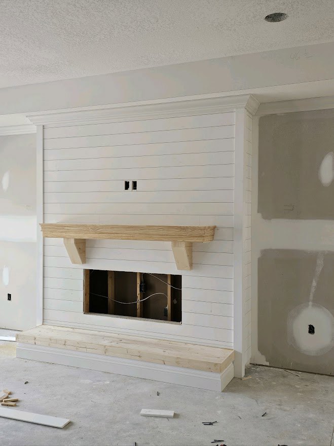 White plaster wall with wood shelf above built-in fireplace, wood paneling detail, white ceiling, exposed wire emerging from wall, black circular object nearby