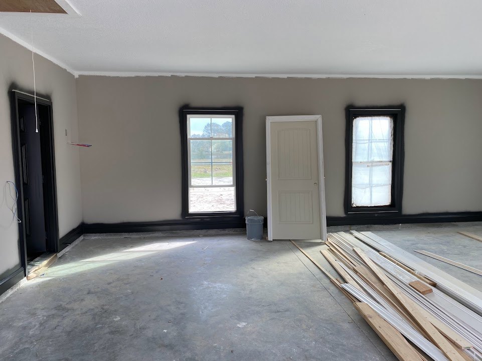 White door with matching frame beside window with sheer white curtain, view of grassy field outside; pile of wood planks stacked on light-colored plaster floor; black door with