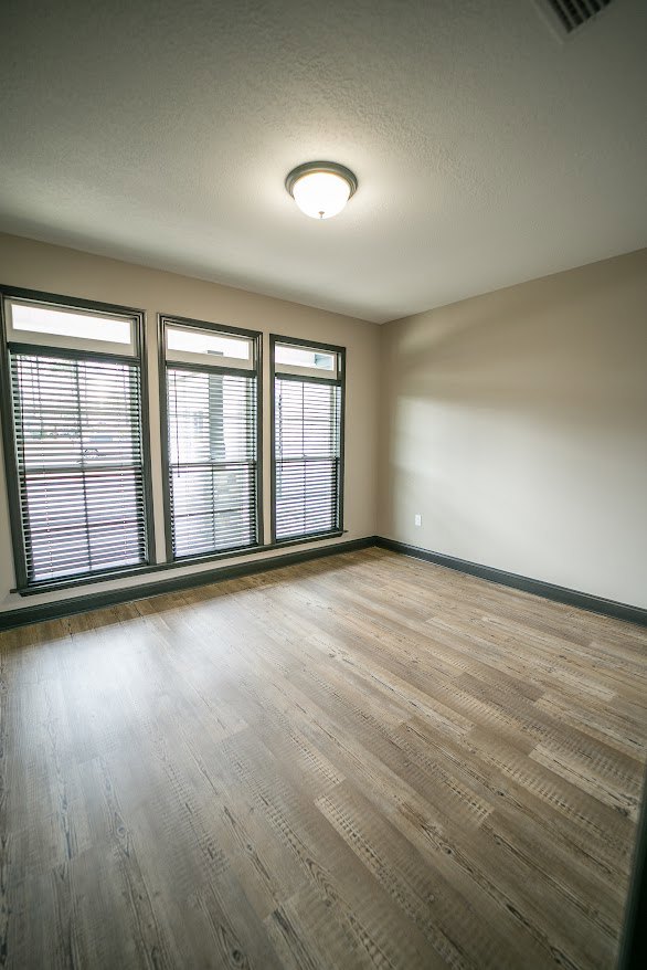 Wood flooring in a bright room with a row of windows fitted with blinds, white walls, and a ceiling-mounted light fixture