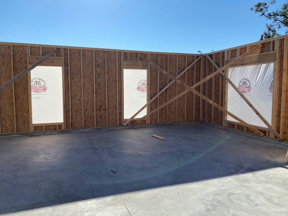 Wood-framed residential structure under construction with exposed beams, white tarps draped over sections, concrete floor, temporary door opening, and tree branches visible in the