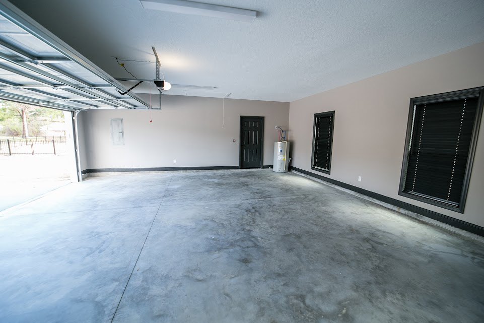 Spacious garage with smooth concrete floor, black overhead door, white side door, and bright natural light illuminating plaster walls and ceiling.