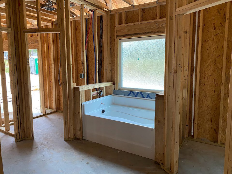 White bathtub set in a wood frame beneath frosted window with blue dot, surrounded by wood walls, exposed beams, and visible white pipe