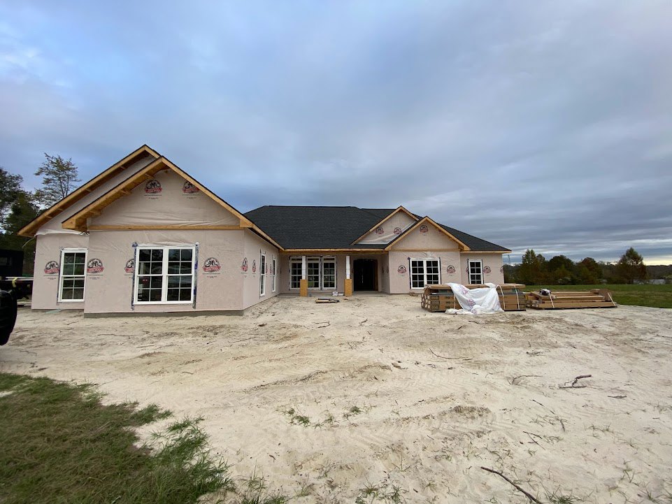 Two-story house under construction with multi-pane windows, unfinished roof, and dirt yard surrounded by scattered building materials