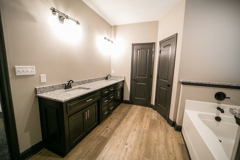 Bathroom featuring a freestanding white bathtub with black faucet, double sink vanity with white countertop, black hardware on doors, wall-mounted light fixture, and light tile