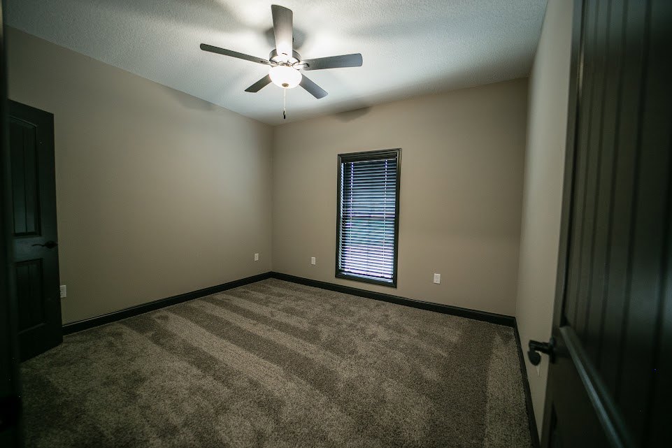 Carpeted room with white walls, ceiling fan with light fixture, window with horizontal blinds, and a door with silver handle and framed artwork.