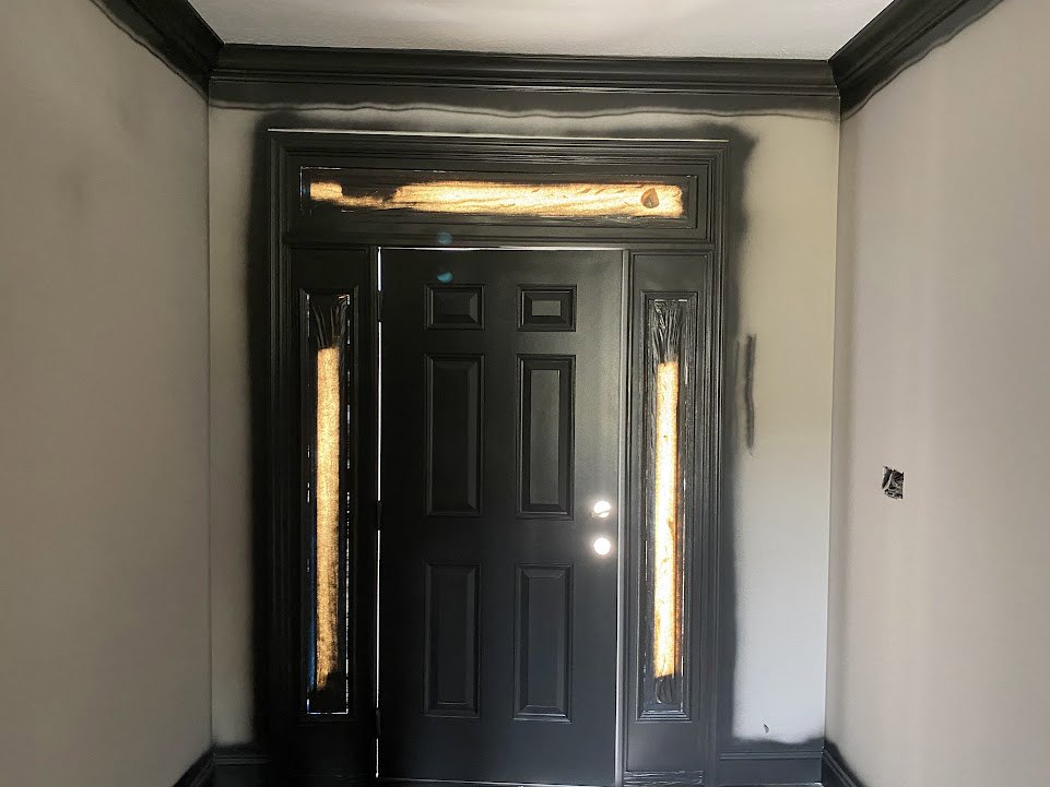 Black painted door with matching black frame, silver door handle, illuminated wall sconce, light-colored walls, and hardwood floor.