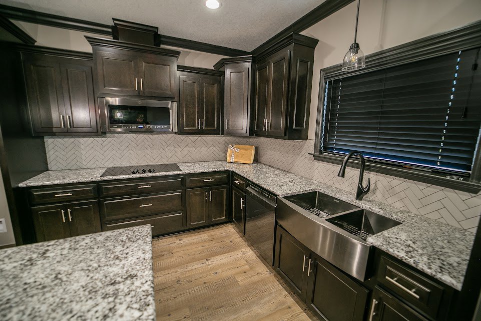 Kitchen with dark wood cabinets, granite countertops, stainless steel sink, microwave, window letting in natural light, and brown carpeted floor