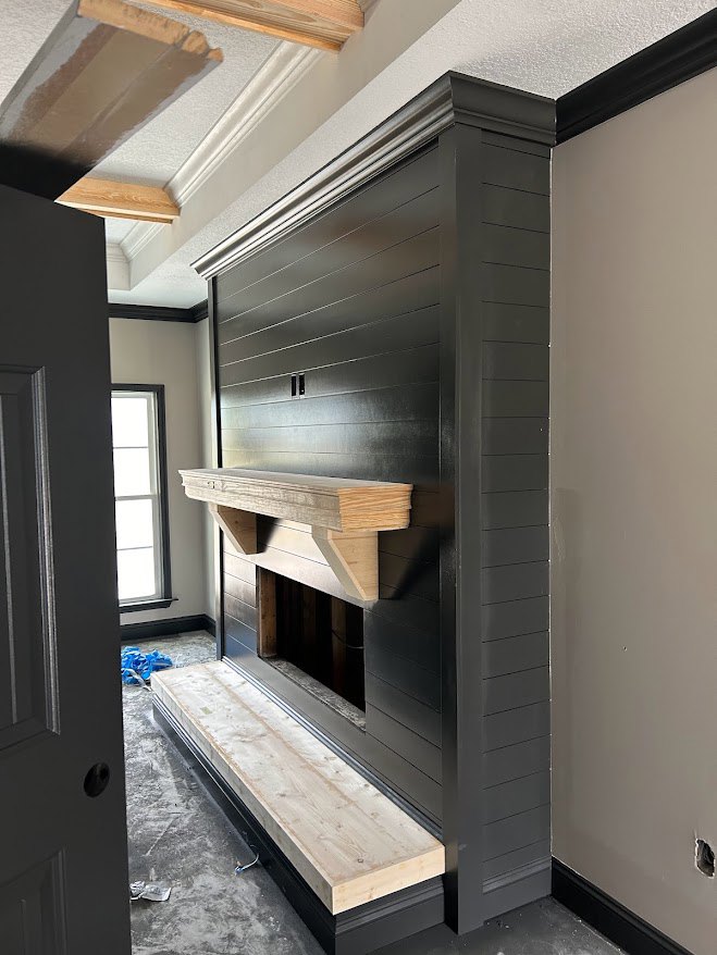 Modern living room with black accent wall, wooden beam above plaster fireplace, built-in wooden bench, light plywood flooring, and white ceiling.