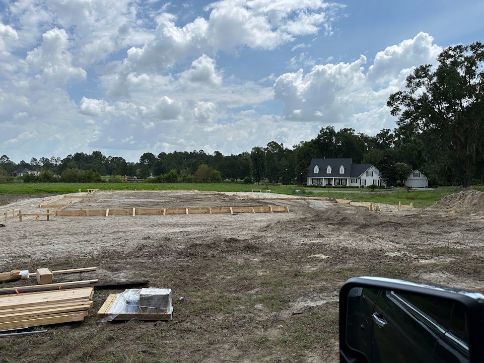 Framed house under construction with black roof, surrounded by grassy field and trees under blue sky; stack of wood planks and plastic-wrapped drain hole in foreground