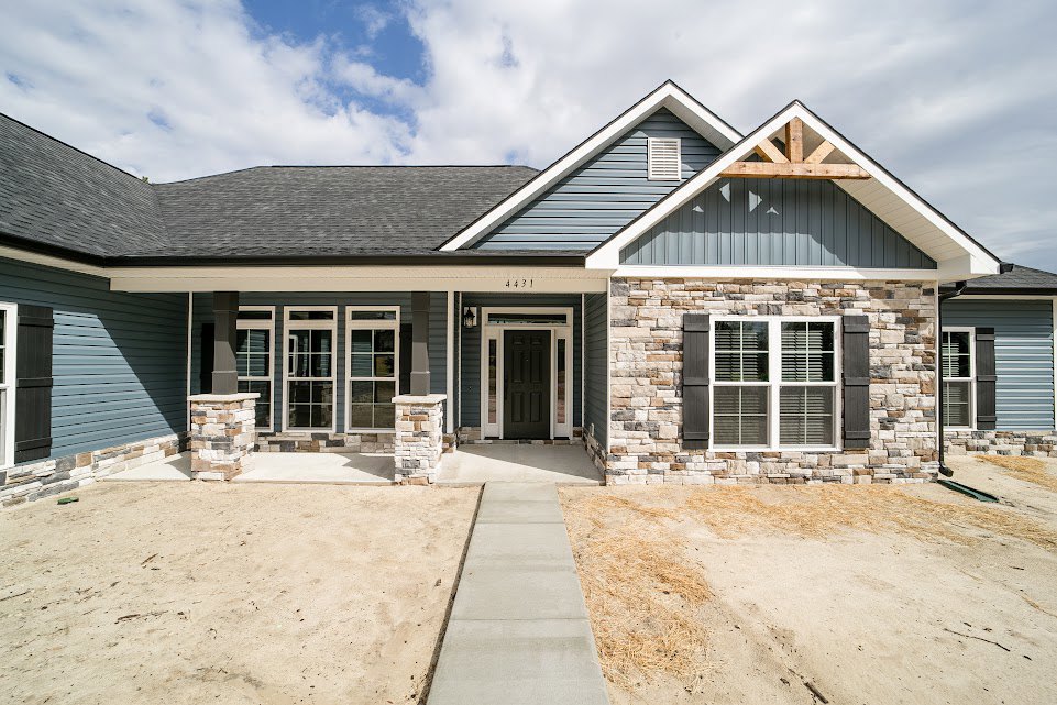 Brick home with white-trimmed black front door, shuttered windows, white window frames, sand and dirt sidewalk, and paved driveway under partly cloudy sky