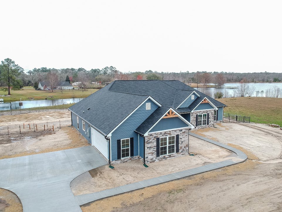 Brick house with blue roof, large windows, concrete driveway with curved edge, green tree in front, river and metal fence in background