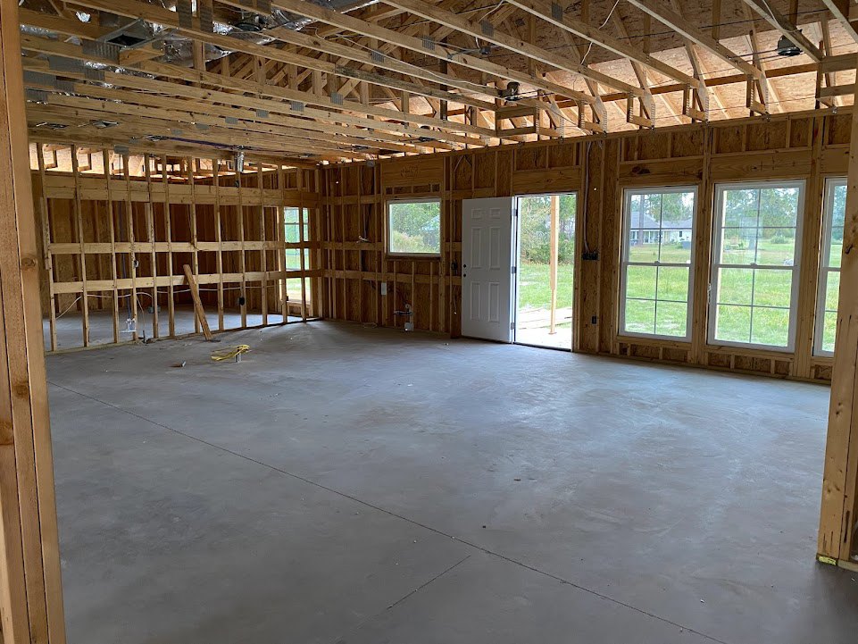 Unfinished room with exposed wooden ceiling beams, concrete floor, white door with metal doorknob, two windows showing grass and neighboring house outside