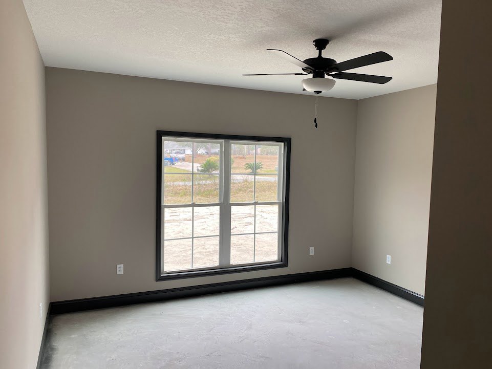 Ceiling fan with light fixture mounted on white plaster ceiling, large window framed in black overlooking fenced grassy yard, smooth white floor, neutral walls