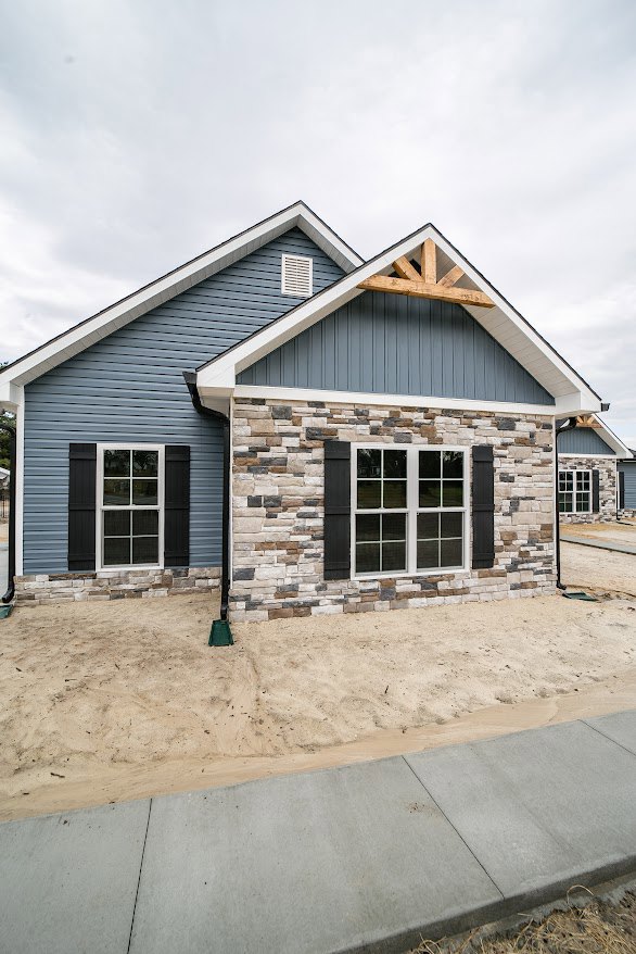 Modern home under construction featuring stone wall, exposed wood beam, white-framed windows, black shutters, sandy ground, and partially finished sidewalk under cloudy sky