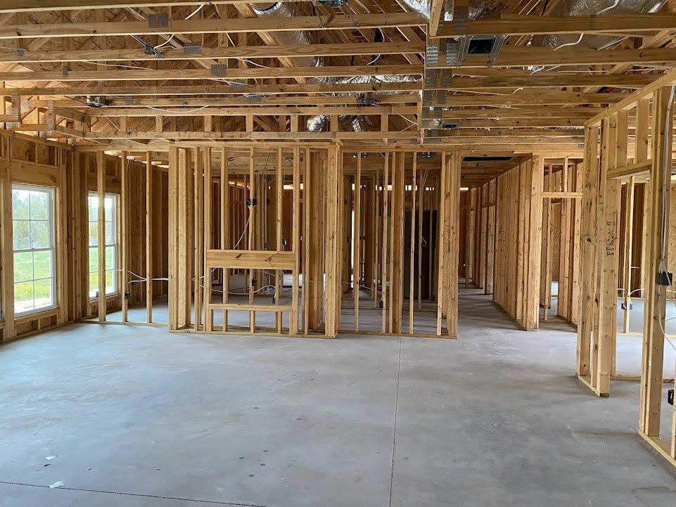 Exposed wood framing and beams over a concrete floor, unfinished ceiling with electrical wiring, large window facing green lawn