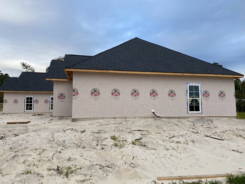 Partially built house with white exterior walls featuring construction logos, white-framed window, dark roof, sandy ground scattered with sticks and grass, cloudy sky overhead